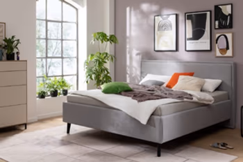 Hier sehen Sie ein Schlafzimmer. Ein Polsterbett mit grauem Stoff und eine Kommode. Unter dem Bett liegt ein heller flacher Teppich.